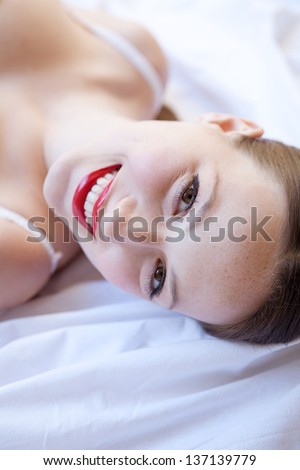 Over head beauty portrait of an attractive young woman laying down on a white bed, wearing white lingerie and glossy red lips, smiling at camera.