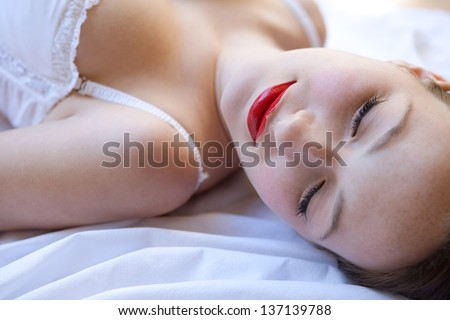 Over head beauty portrait of an attractive young woman laying down on a bed, wearing white lingerie and glossy red lips, looking down.