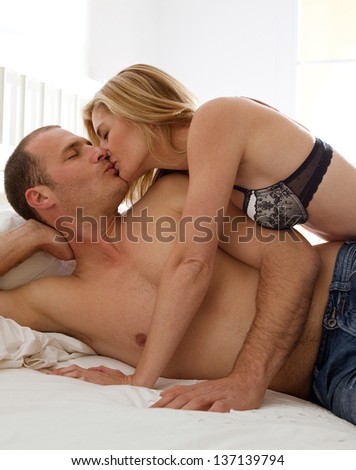 Attractive sexy couple being intimate and kissing while laying down on a white bed in a home bedroom.