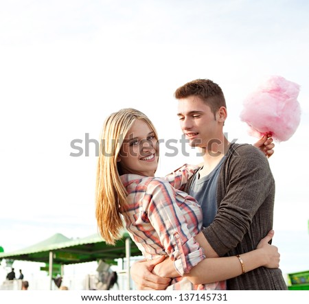 Attractive young couple hugging and having fun in an amusement park arcade during a sunny day, holding a cotton sugar sweet and turning to the camera smiling.