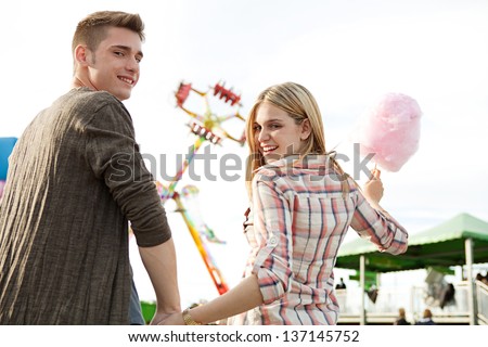 Attractive young couple hugging and having fun in an amusement park arcade, holding a cotton candy sweet and turning to the camera smiling.