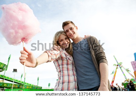 Attractive young couple holding each other and having fun in an attractions park arcade, holding a cotton candy sweet and smiling.