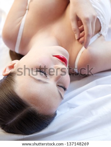 Over head beauty portrait of an attractive young woman laying down on a white bed, wearing white lingerie and glossy red lips.
