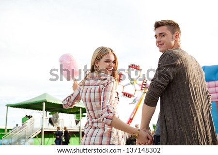 Attractive young couple holding hands and having fun in an attractions park arcade, holding a cotton candy sweet and turning to the camera smiling.