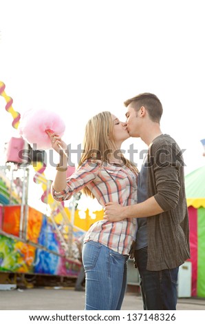 Young couple having fun in a colorful attractions park arcade, holding cotton candy and kissing spontaneously.