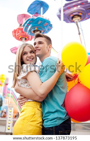 Young couple hugging and holding each other while visiting an attractions park arcade with lights and rides in the background.