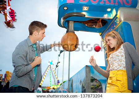 Young teenage couple visiting an attractions park arcade, playing punch boxing game during early evening with lights and rides in the background.