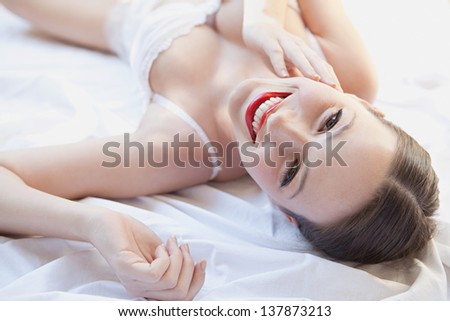 Over head beauty portrait of a young woman laying down in bed wearing white lingerie and red lips, smiling at camera.