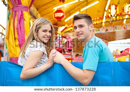 Teenage couple visiting a fun fair amusement arcade, sitting near a colorful ride with lights, holding hands and smiling.