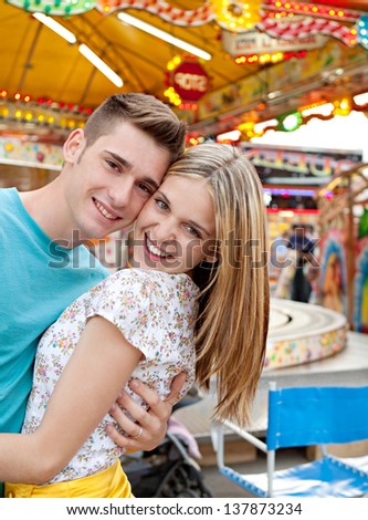 Joyful young couple holding each other while visiting an attractions park arcade with lights and colorful rides in the background, close up portrait.