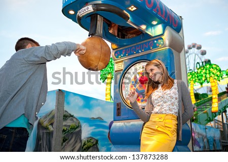 Young teenage couple visiting an attractions park arcade, playing punch boxing game during early evening with lights and rides in the background.