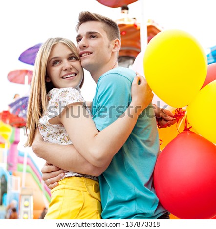 Young couple hugging and holding each other while visiting an attractions park arcade with lights and rides in the background.