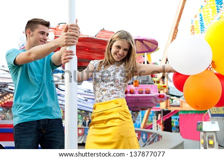 Young couple of teenagers visiting a fun fair ground with rides and lights around them, holding balloons, being playful and joyful during a sunny day.