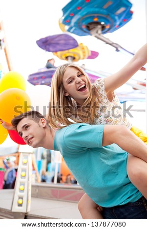 Joyful young couple being playful while visiting an attractions park arcade with rides, with young man giving girl a piggy bag and having fun.
