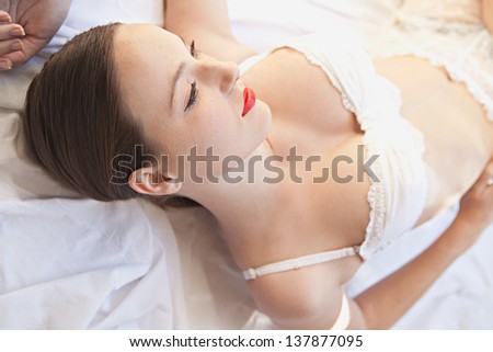 Over head beauty portrait of a young woman peacefully laying down in bed wearing white lingerie and red lips.