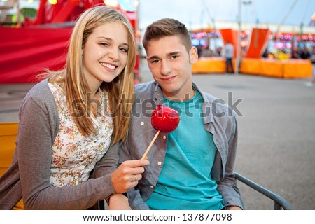 Teenage couple visiting a fun fair amusement arcade, sitting near a colorful ride with lights, holding a caramel red sugar apple and smiling.