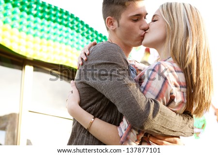 Close up of a young couple visiting an amusement park arcade kissing during a sunny day with a box office and rides in the background.