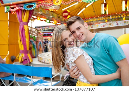 Joyful young couple holding each other while visiting an attractions park arcade with lights and colorful rides in the background, close up portrait.