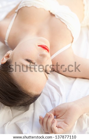 Over head close up beauty portrait of a young woman peacefully laying down in bed wearing white lingerie and red lips.
