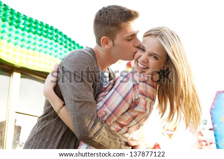 Close up of a young couple visiting an amusement park arcade with man kissing the girl on the cheek during a sunny day.