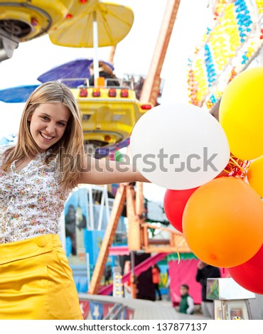 Portrait of a teenage girl visiting a fun fair ground with rides and lights around her, holding balloons being playful and joyful during a fun day.