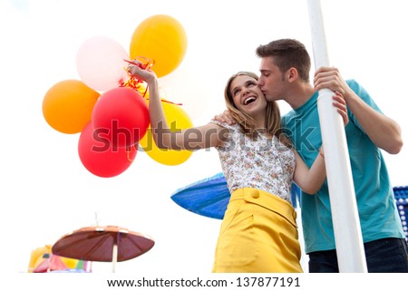 Young couple of teenagers visiting a fun fair ground with rides around them, holding balloons, being joyful and kissing during a sunny day.