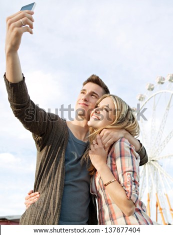 Young couple posing together at an attractions park arcade and using their smartphone to take a picture of themselves smiling.
