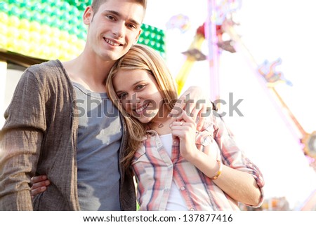Joyful young couple holding each other while visiting an attractions park arcade with a ticket office and rides in the background, close up portrait.