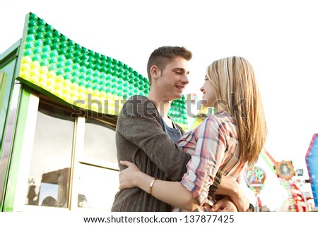 Young couple hugging and holding each other while visiting an attractions park arcade with a ticket office and rides in the background.