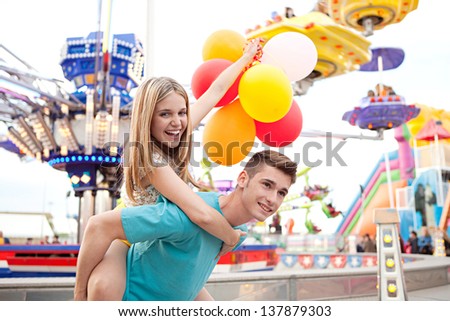 Joyful young couple being playful while visiting an attractions park arcade with rides, with young man giving girl a piggy bag and having fun.