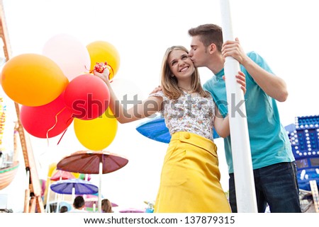 Young couple of teenagers visiting a fun fair ground with rides and lights around them, holding balloons, being joyful and kissing during a sunny day.