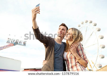 Young couple posing together at an attractions park arcade and using their smartphone to take a picture of themselves smiling.