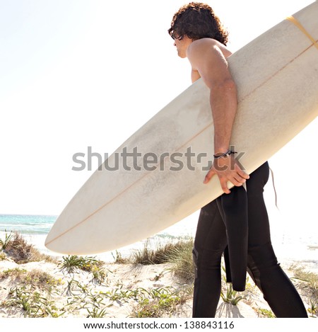 Young surfer walking towards the shore of a white sand beach carrying his surfing board and wearing a neoprene rubber suit during a sunny day with a blue sky.