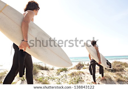 Two Young Surfer Friends Walking Towards The Shore Of A White Sand Beach Carrying Their Surfing Boards And Wearing A Neoprene Rubber Suit During A Sunny Day With A Blue Sky.