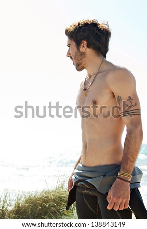 Young attractive surfer sports man wearing a neoprene rubber diving suit and looking at the horizon while standing on a beach during a sunny day.