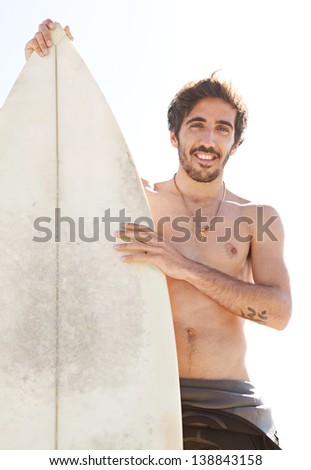 Young surfer sports man standing on a beach while wearing a neoprene rubber surfing suit during a sunny day, holding a surfing board upright and smiling at camera.