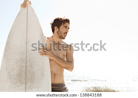 Young surfer sports man standing on a beach while wearing a neoprene rubber surfing suit during a sunny day, being thoughtful and holding a surfing board upright.