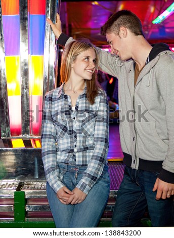 Attractive young couple visiting an amusement park arcade standing by a bouncy cars ride, talking and having fun during an exciting night out.