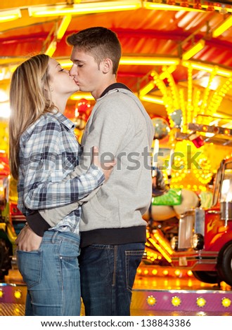 Profile portrait of an attractive young couple visiting an amusement park arcade with a colorful carousel, holding each other and kissing during a fun night out.