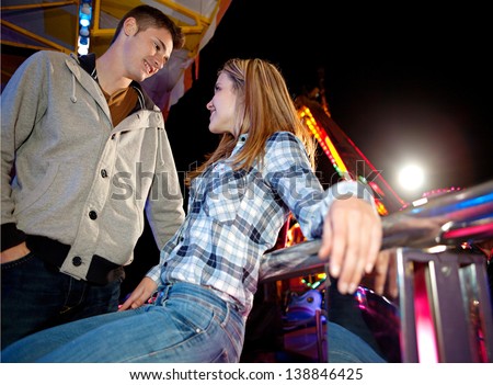Young teenager couple visiting a funfair ground arcade and talking while sitting down near attractions rides at night time.