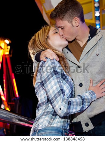 Young teenager couple visiting a funfair ground arcade and kissing on the lips while sitting down near attractions rides with colorful lights at night time.