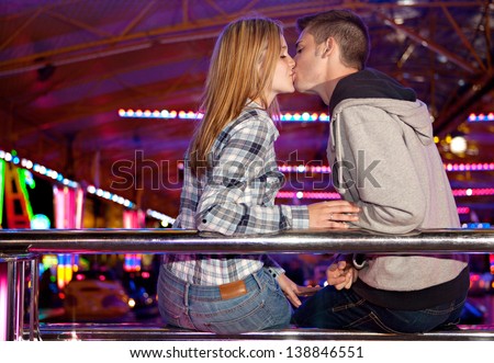 Young couple sitting by a bouncy cars ride while visiting an attractions park ground, kissing on the lips with colorful lights in the background at night time.