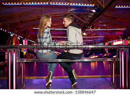 Young couple sitting by a bouncy cars ride while visiting an attractions park ground, turning to camera and smiling with colorful lights in the background at night time.