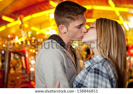 Close up side portrait of an attractive young couple visiting an amusement park arcade with a colorful carousel, holding each other and kissing during a fun night out.