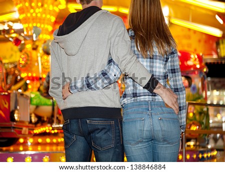 Rear middle section view of a young couple standing by an amusement park arcade carousel ride with colorful lights at night, and holding each other.