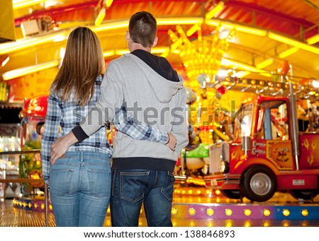Rear view of a young couple standing by an amusement park arcade carousel ride with colorful lights at night, and holding each other.