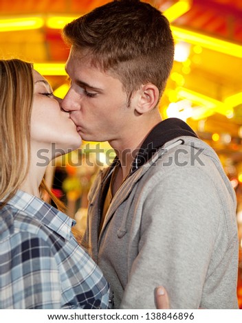 Close up side portrait of an attractive young couple visiting an amusement park arcade with a colorful carousel, holding each other and kissing during a fun night out.