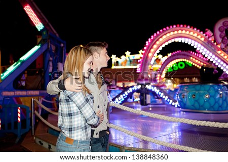 Side portrait view of a young couple visiting an attractions park arcade and getting excited while looking at a colorful ride at night time.