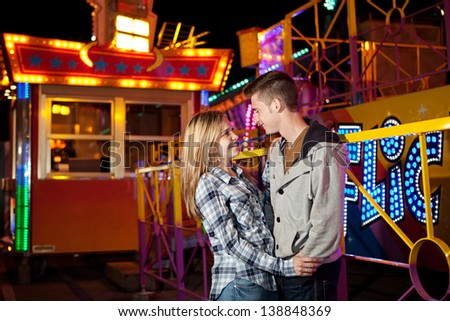 Portrait of a young couple visiting an attractions park arcade being romantic and holding each other with rides and lights in the background during night time.