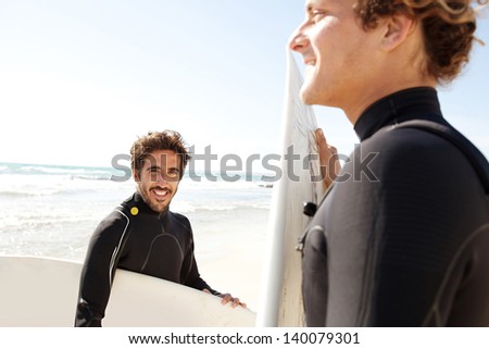Two young surfers sports men wearing surfing neoprene waterproof suits and carrying their surfing boards on a white sand beach with a blue sky in the background during a sunny day.